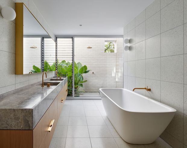 Bright contemporary bathroom featuring a freestanding white bathtub, wall-mounted brass tapware, a timber vanity with a thick grey stone benchtop, and a large mirror. Pale tiled walls and floor create a clean, minimal setting, while full-height glazing and louvred windows bring in greenery and natural light.