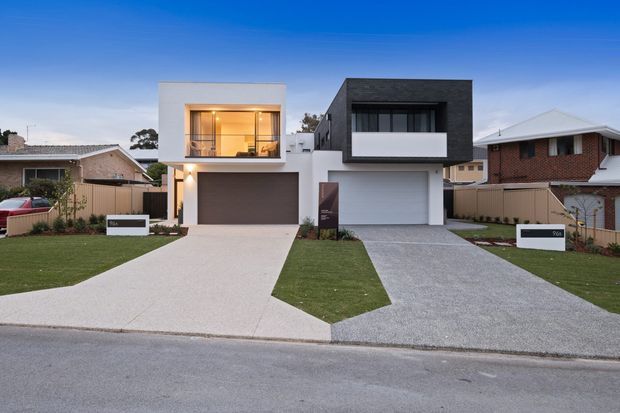 Street-facing view of two adjacent contemporary two-storey homes with boxy forms, flat rooflines, contrasting light and dark rendered facades, large upper-level glazing, integrated garages, and separate concrete driveways and lawns. Traditional brick suburban homes are visible on either side.