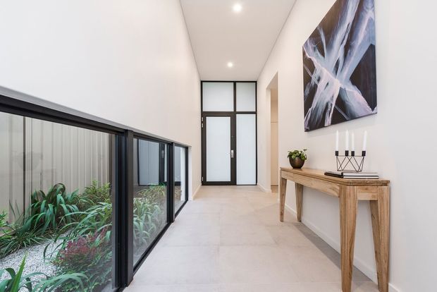 Bright contemporary hallway with a long internal glass-sided courtyard or garden view, pale tiled flooring, white walls, black-framed glazed doors, and a slim rustic timber console styled with a plant, candles, and abstract artwork.