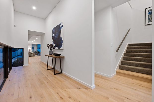 Bright modern residential entry hall with pale timber flooring, white walls and ceiling, recessed downlights, a slim console table with artwork, and an adjacent carpeted staircase with a simple timber handrail. Glazed doors and a living area are visible deeper in the home.