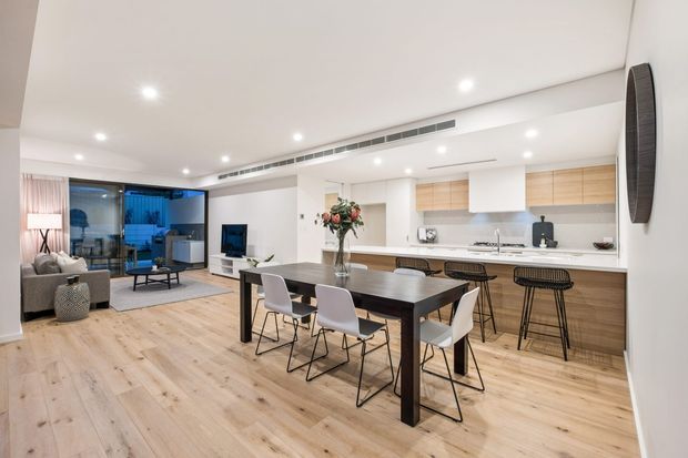 Open-plan contemporary living, dining and kitchen interior with light timber flooring, a dark dining table, a long kitchen island with bar seating, pale cabinetry, recessed ceiling lights and large glazed doors opening to an outdoor area. The living zone includes a sofa, coffee table and television, while the kitchen features a white benchtop and timber cabinetry.