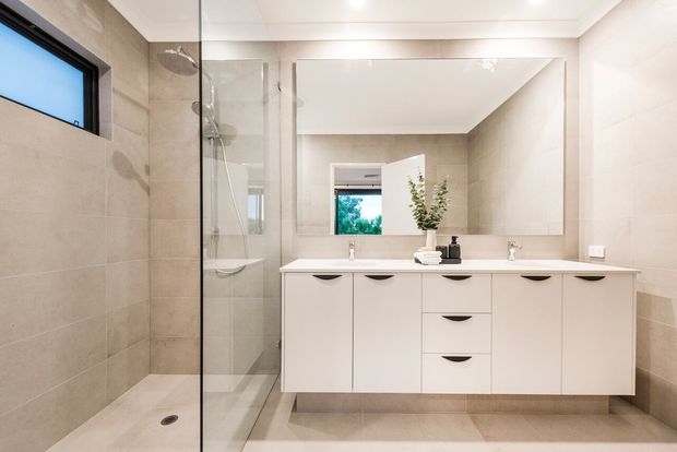 Bright contemporary bathroom with a long floating white vanity, integrated basins, large wall mirror, frameless glass walk-in shower, and warm beige tiled walls and floor. A small high-set window and recessed ceiling lighting provide natural and artificial illumination.