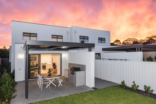 A contemporary two-storey white-rendered home viewed from the rear, with a covered outdoor entertaining patio, outdoor kitchen or barbecue bench, dining setting, lawn and a high white privacy fence. Large glazed doors connect the patio to the interior, while a dramatic pink-orange sunset forms the background.