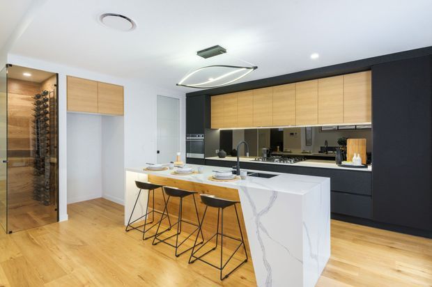 Contemporary open-plan kitchen with a large white marble-look island and integrated breakfast bar, black lower cabinetry and feature wall, warm light timber upper cabinets and flooring, and sleek modern pendant lighting. A built-in wine storage area and pantry joinery are visible to the left.