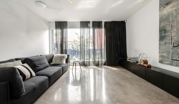 Contemporary living room with a large charcoal sectional sofa, black low-profile built-in media cabinetry, floor-to-ceiling glazing with sheer and blackout curtains, polished light-grey concrete flooring, and a large abstract wall artwork. The room is bright and open with a restrained monochromatic palette.