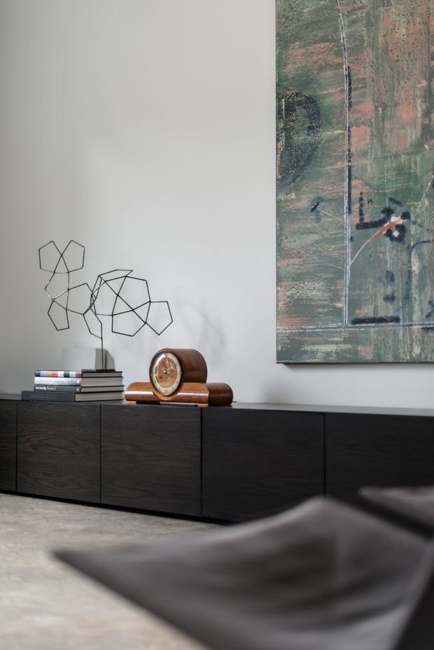 A refined living room vignette featuring a long, low dark timber sideboard against a pale wall. The sideboard is styled with stacked books, a geometric wire sculpture, and a vintage wooden mantel clock, while a large abstract artwork hangs above. A dark leather lounge chair is partially visible in the foreground, and the room has a polished concrete floor.