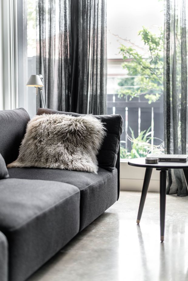 A softly lit living room corner featuring a dark charcoal sectional sofa, a shaggy neutral throw pillow, sheer charcoal curtains, a slim floor lamp, and a small round side table with books. Large windows overlook greenery and a fenced outdoor area, while the polished light-grey concrete floor reflects the daylight.