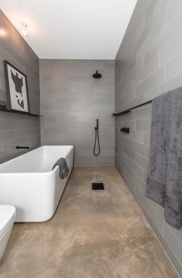 A contemporary bathroom featuring a freestanding white bathtub, open walk-in shower, grey horizontal wall tiles, a warm concrete-look floor, black shower and towel hardware, and simple black wall shelving. The space has a clean, restrained industrial-modern character with a framed monochrome artwork and grey towels.