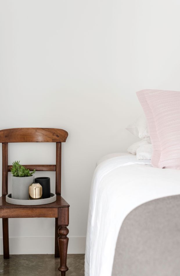 A softly lit bedroom vignette featuring a white bed with layered white bedding and a pale pink textured cushion, beside a traditional dark timber chair used as a bedside table. A round tray with a potted plant and small decorative vessels sits on the chair. The room has warm grey polished concrete flooring and plain off-white walls, creating a calm, minimal, contemporary look with rustic timber character.