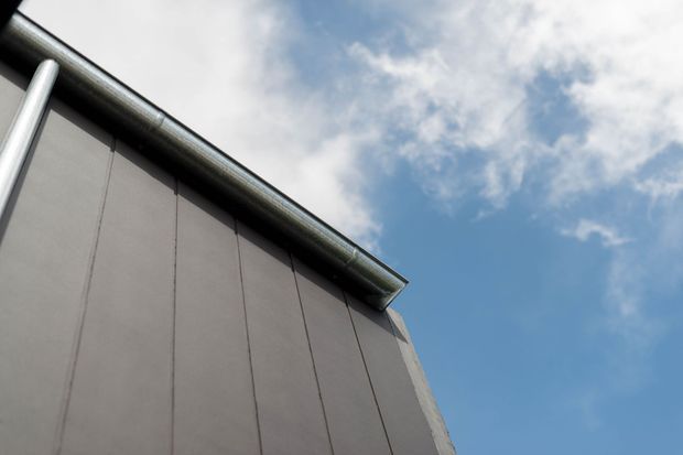 Close-up upward view of a contemporary building facade with large smooth grey vertical panels and a dark metal rain gutter or roof edge. A tubular metal downpipe is visible along the left side, set against a partly cloudy blue sky.