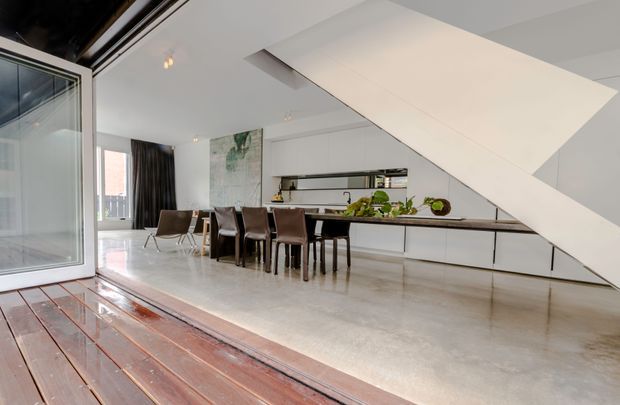 Bright contemporary open-plan kitchen and dining area viewed from a timber deck through large glazed doors. The space features a long white island with integrated cabinetry, dark dining chairs, polished concrete flooring, white walls, and a dramatic angular stair or ceiling structure sweeping across the room. Indoor plants and a partially visible living area add warmth.