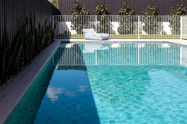 A contemporary residential swimming pool in a landscaped courtyard, with turquoise mosaic-style tile lining, dark vertical cladding walls, a white metal balustrade, and sculptural white outdoor seating. Tall narrow plants line the left boundary, while climbing greenery softens the rear wall.