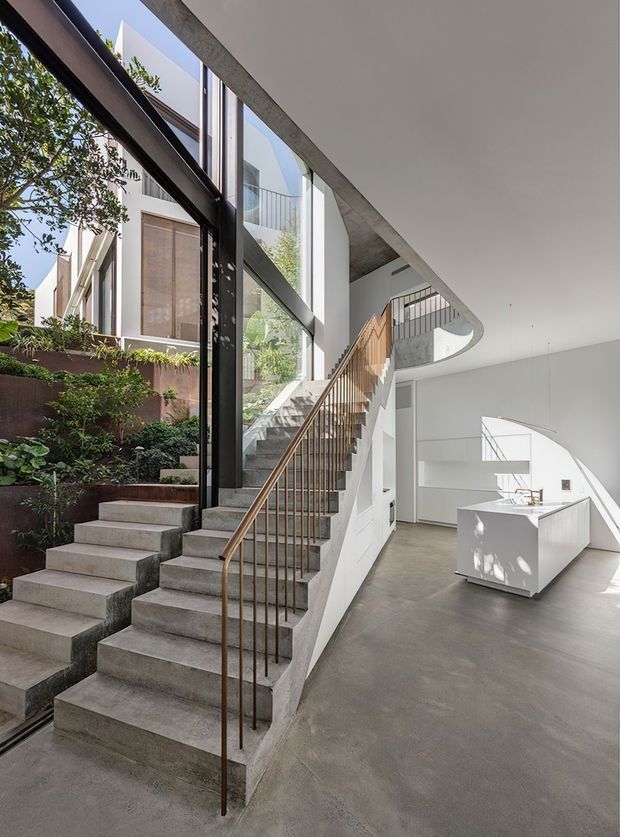 A bright, double-height modern interior combining a sculptural concrete stair with slender metal balustrade, black-framed floor-to-ceiling glazing, and an open white kitchen. The polished concrete floor and exposed concrete ceiling edge contrast with crisp white walls and warm timber stair detailing. A planted courtyard is visible through the glazing, bringing greenery and daylight into the space.
