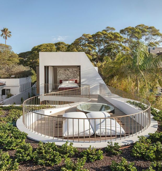 A striking rooftop terrace with a sculptural white architectural form, curved metal balustrades, integrated outdoor seating and a circular recessed feature. A bedroom opens directly onto the terrace, with lush landscaped planting and mature trees surrounding the property.