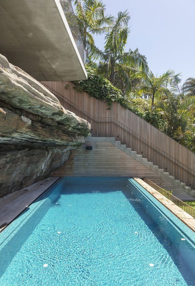 A narrow rectangular swimming pool occupies the foreground beside a dramatic natural rock outcrop. Timber-clad retaining walls and integrated stairs rise behind the pool, with dense tropical palms surrounding the outdoor space. A projecting concrete slab forms an overhead architectural element at the upper left. The scene is bright, warm, and resort-like, with strong integration of landscape and architecture.