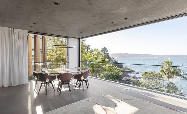 A bright contemporary dining area with a dark timber dining table and sculptural upholstered chairs, positioned beside expansive floor-to-ceiling glazing overlooking a tropical coastal bay. The room features a smooth concrete ceiling and floor, sheer white curtains, minimalist recessed lighting, and an open, airy connection to the landscape.