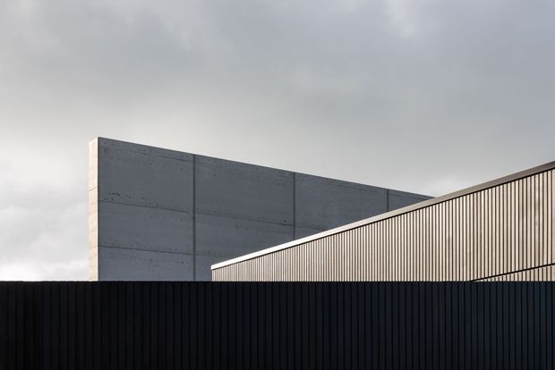 Minimalist architectural exterior showing a dark vertical-slat fence in the foreground, a large exposed concrete wall behind it, and a sharply sloping light-toned metal-clad roof or facade plane. The overcast sky and restrained monochrome palette create a stark, contemporary composition.
