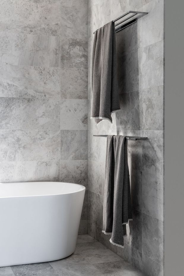 A close-up of a refined contemporary bathroom featuring large-format grey stone-look wall and floor tiles, a white freestanding bathtub, and minimalist dark metal towel rails holding textured grey towels.