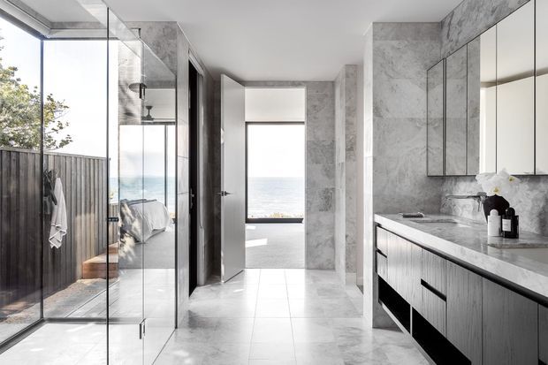 A bright, contemporary bathroom with a frameless glass shower on the left, a long dark timber vanity with a pale stone benchtop on the right, large grey-white marble-look wall and floor tiles, and a freestanding bathtub visible through the central doorway. Full-height glazing opens the space toward an ocean view, while a mirrored wall expands the sense of depth.