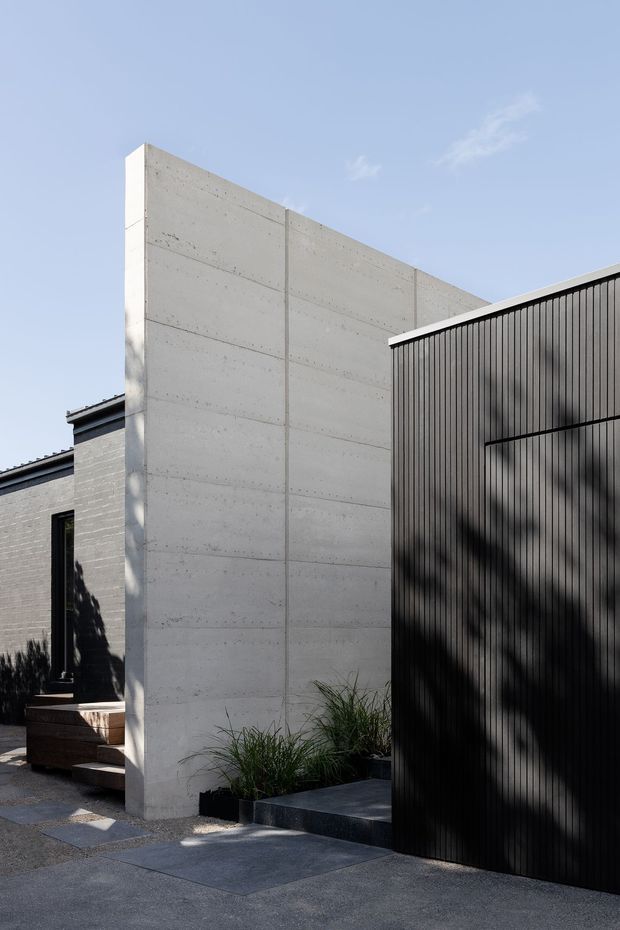A modern residential exterior featuring a tall smooth board-formed concrete wall, dark vertically ribbed metal cladding, and a low grey masonry building. Paved stepping stones and minimalist ornamental grasses sit in front, with strong sunlight and shadows creating a restrained, sculptural composition.