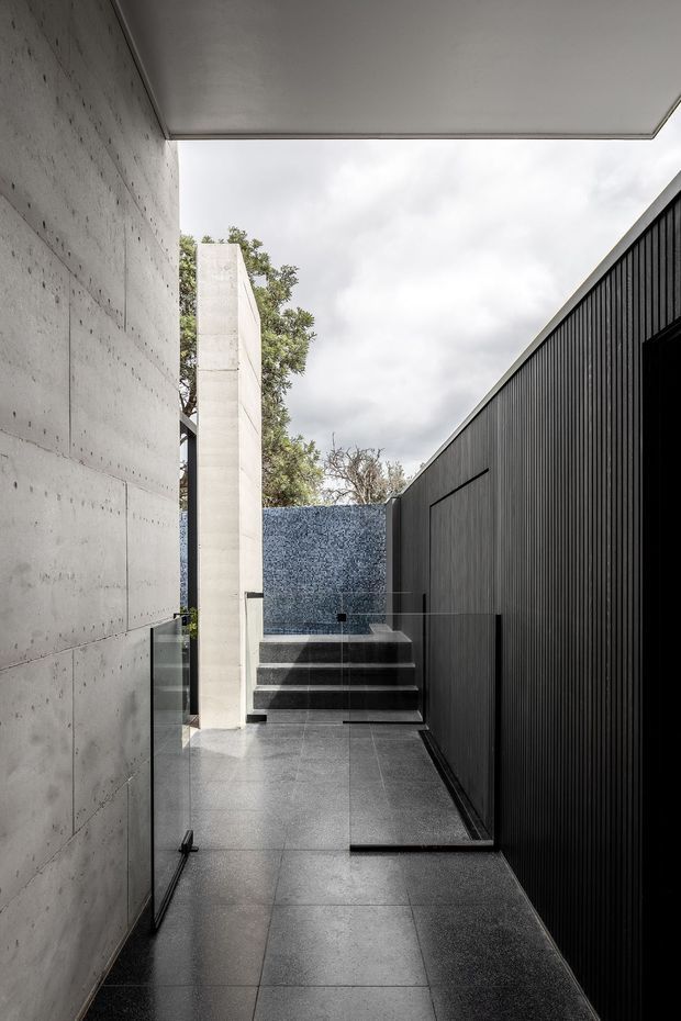 A narrow, contemporary exterior passage with large-format dark grey tiled paving, raw concrete walls, black vertical metal cladding, glass balustrades, and a short flight of steps leading toward a blue-grey tiled feature wall. The partially covered walkway has a strong geometric, minimalist architectural character.