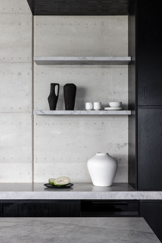 Close-up of a refined kitchen or entertaining joinery wall featuring open marble shelves, a pale concrete backsplash, dark timber cabinetry, and a marble benchtop. Minimal ceramic vessels and tableware are arranged on the shelving and counter.
