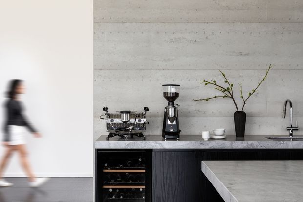 Contemporary kitchen and coffee station with a textured pale concrete wall, dark timber cabinetry, stone benchtops, integrated wine storage, espresso machine and grinder. A blurred person passes through the adjacent open area.