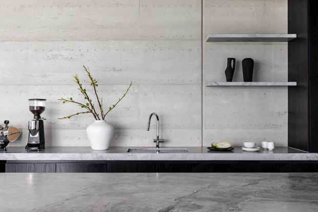 Minimalist contemporary kitchen with a long grey stone island in the foreground, a concrete-look wall and splashback, integrated sink and chrome gooseneck tap, dark textured cabinetry, and open shelves displaying simple black ceramics and coffee-making equipment. The restrained monochrome palette and clean lines create a refined, industrial-leaning interior.