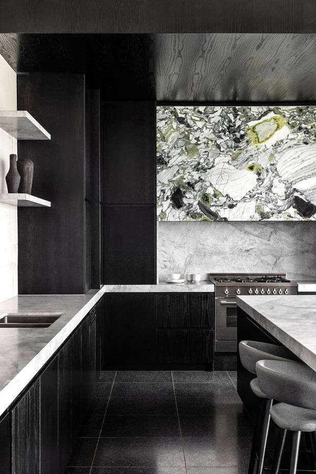 A dramatic contemporary kitchen with black vertically grained cabinetry, dark tiled flooring, pale stone benchtops, a large veined stone splashback or feature panel, integrated shelving, and stainless-steel cooking appliances. A central island with grey upholstered bar stools occupies the foreground. The palette is monochromatic charcoal, white, grey, and natural stone with strong veining.