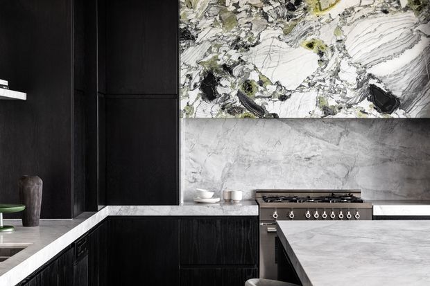 A dramatic contemporary kitchen with matte black timber-look cabinetry, pale stone benchtops, a large grey-veined island, and a striking bookmatched-style marble or stone feature splashback above a freestanding range. The palette is monochromatic with strong natural stone veining and refined dark joinery.
