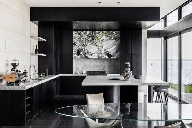 A sophisticated contemporary kitchen with black timber cabinetry, pale stone benchtops and splashback, a central island, integrated appliances, open shelving, and a glass dining table. Full-height glazing on the right opens to a bright waterfront view, while monochromatic artwork and sculptural accessories add visual interest.