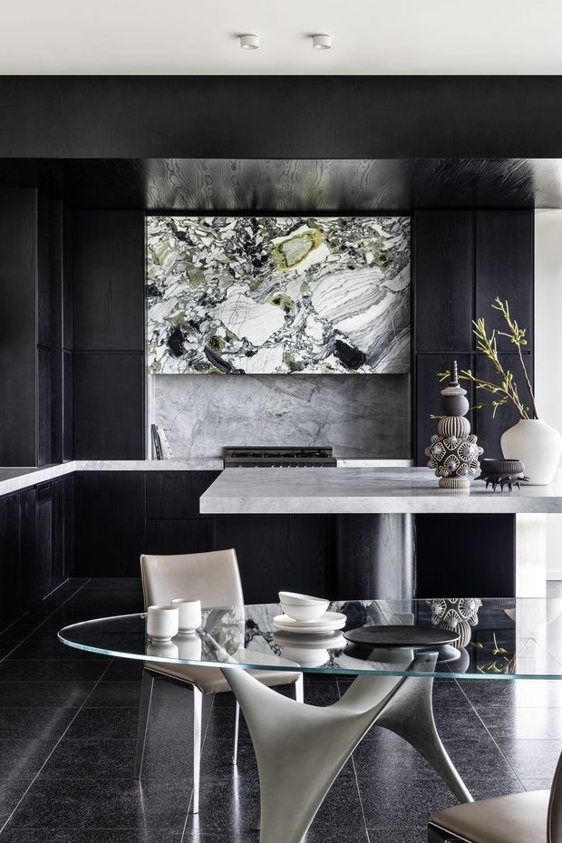 A dramatic, high-end open-plan kitchen and dining space featuring dark timber cabinetry, a large pale stone island, a striking veined stone splashback, and polished dark tiled flooring. A sculptural glass dining table with sculptural chairs occupies the foreground, complemented by monochromatic decorative objects and contemporary lighting.