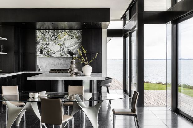 A sophisticated open-plan kitchen and dining area with black cabinetry, a pale stone island and splashback, and a glass dining table surrounded by sculptural chairs. Floor-to-ceiling black-framed glazing opens toward a waterfront deck and lawn, creating a bright, high-contrast interior with polished dark flooring and contemporary decorative styling.