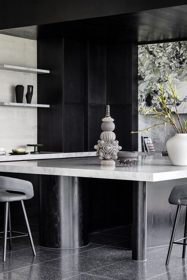 A sophisticated contemporary kitchen and bar area with a dark timber or black-stained cabinetry wall, pale stone benchtop and island, dramatic veined stone splashback, and dark polished flooring. Sculptural decorative objects, open shelving, stools, and a potted branch add visual interest to the monochromatic scheme.