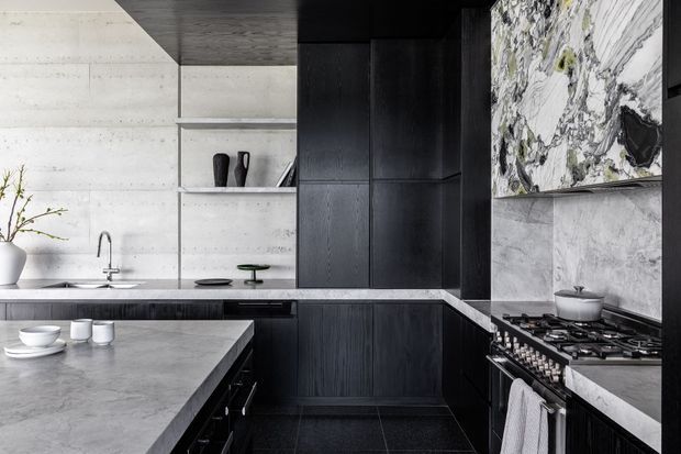 Contemporary kitchen featuring dark timber cabinetry, pale stone benchtops, a dramatic veined marble splashback, and a large island. Concrete-look wall panels, integrated shelving, black appliances, and minimal styling create a refined monochromatic interior.
