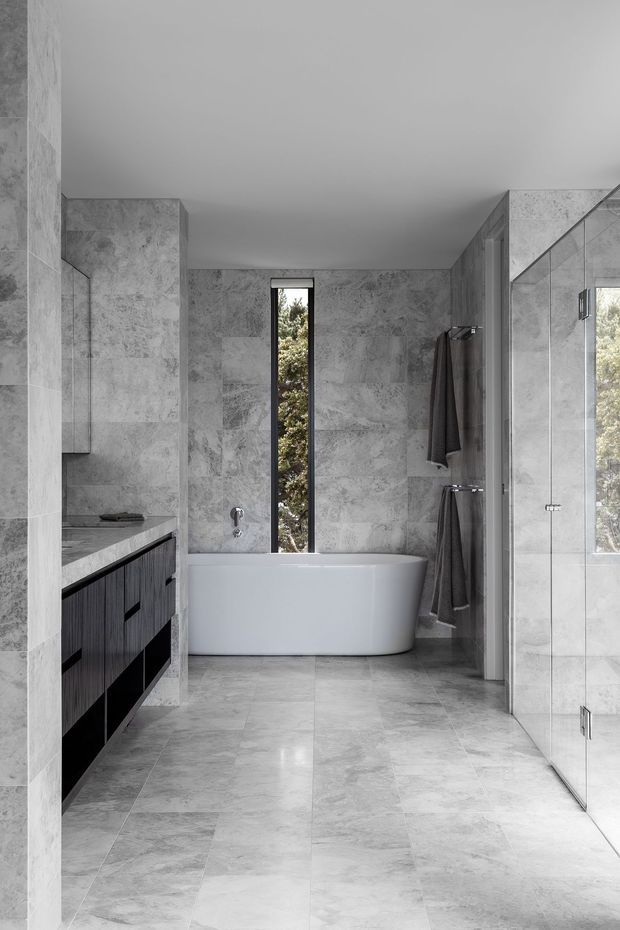 A refined contemporary bathroom clad in large-format grey stone-look tiles, featuring a freestanding white bathtub beneath a narrow vertical window, a dark wood vanity with stone benchtop, and a frameless glass shower enclosure. The monochromatic palette and clean-lined built-in joinery create a calm, luxurious atmosphere.
