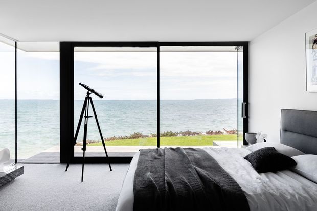Bright contemporary bedroom with a large floor-to-ceiling black-framed glazed wall overlooking the ocean. The room contains a bed with white bedding, a dark charcoal throw and pillows, a grey upholstered headboard, light carpet, and a telescope positioned toward the coastal view.
