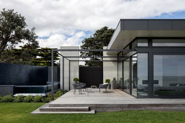 Contemporary single-storey home with a flat roof, dark metal-framed floor-to-ceiling glazing, pale stone or concrete wall panels, and a raised timber-look deck. The outdoor terrace includes minimalist seating and a dark built-in outdoor feature, with landscaped lawn and mature trees surrounding the structure. An ocean or expansive water view is visible through the glazing.