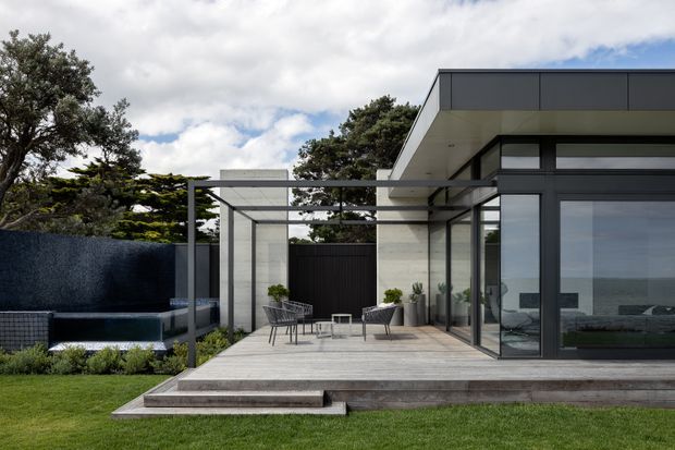 Contemporary single-storey home exterior with a generous timber deck and terrace framed by dark metal posts and beams. Floor-to-ceiling glazing opens onto the deck, with a flat roof, pale masonry walls, dark cladding, and a landscaped lawn. A partially visible outdoor seating and pool area sits to the left, while the glazing reflects a coastal horizon.