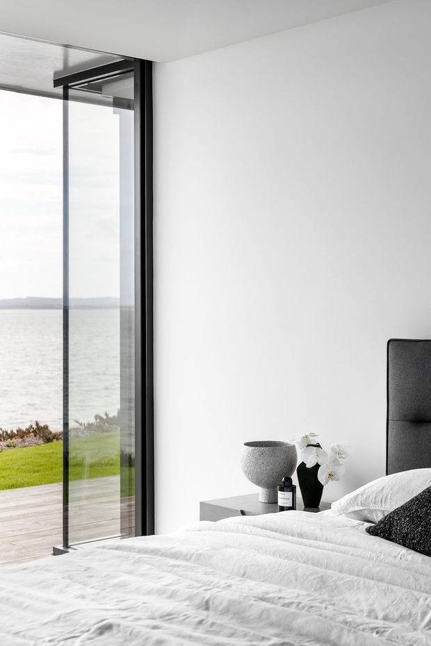 Bright minimalist bedroom with a large frameless glazed opening overlooking a waterfront landscape. The room features white walls and bedding, a dark upholstered bedhead, a slim floating bedside table, and restrained black-and-white accessories. Black-framed glazing and a seamless indoor-outdoor connection create a calm, contemporary setting.
