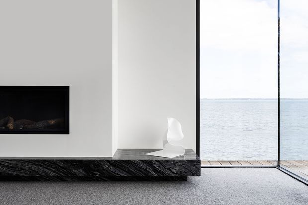 Minimalist contemporary interior with a white feature wall and recessed black fireplace, a dramatic dark veined stone hearth or ledge, grey carpet, and a sculptural white chair. Full-height black-framed glazing opens the room visually toward a calm waterfront view and a narrow timber deck.