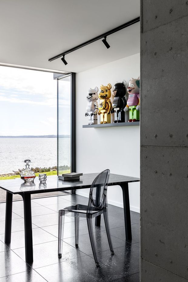 A contemporary dining/work area beside full-height glazing overlooking a calm waterfront. The room features a dark stone or metal rectangular table, transparent acrylic dining chair, polished dark tiled floor, white walls, exposed concrete wall construction, black track lighting, and a wall shelf displaying collectible figures.