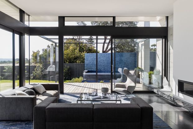 A bright contemporary living room with floor-to-ceiling black-framed glazing opening onto a timber deck and landscaped outdoor area. The room features a large dark sectional sofa, sculptural lounge chairs, a glass coffee table, polished dark stone or tile flooring, and a minimalist built-in fireplace. A blue tiled or textured outdoor wall is visible beyond the glazing.