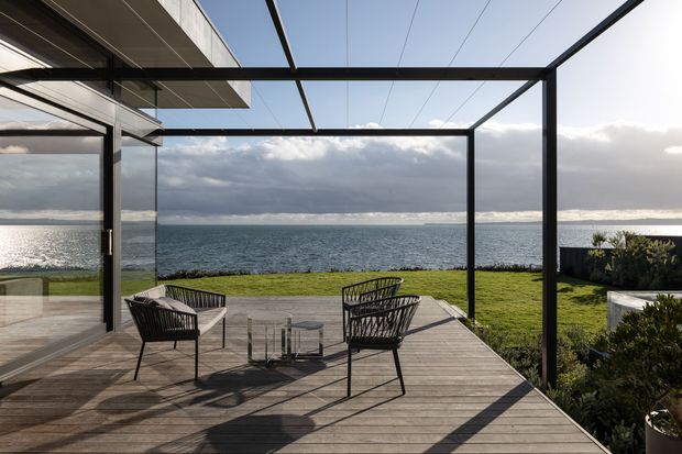 A contemporary waterfront outdoor terrace with a timber deck, minimalist black-framed glass walls and an open pergola structure. Two woven lounge chairs and a small table face the ocean, with a lawn and low coastal planting beyond. Dramatic cloud-filtered sunlight creates strong shadows across the deck.