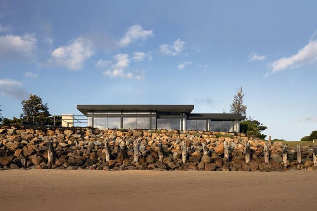 A low-slung contemporary house viewed from across a sandy beach, with extensive floor-to-ceiling glazing, a broad flat projecting roof, dark structural framing, and a rugged stone retaining wall in the foreground.