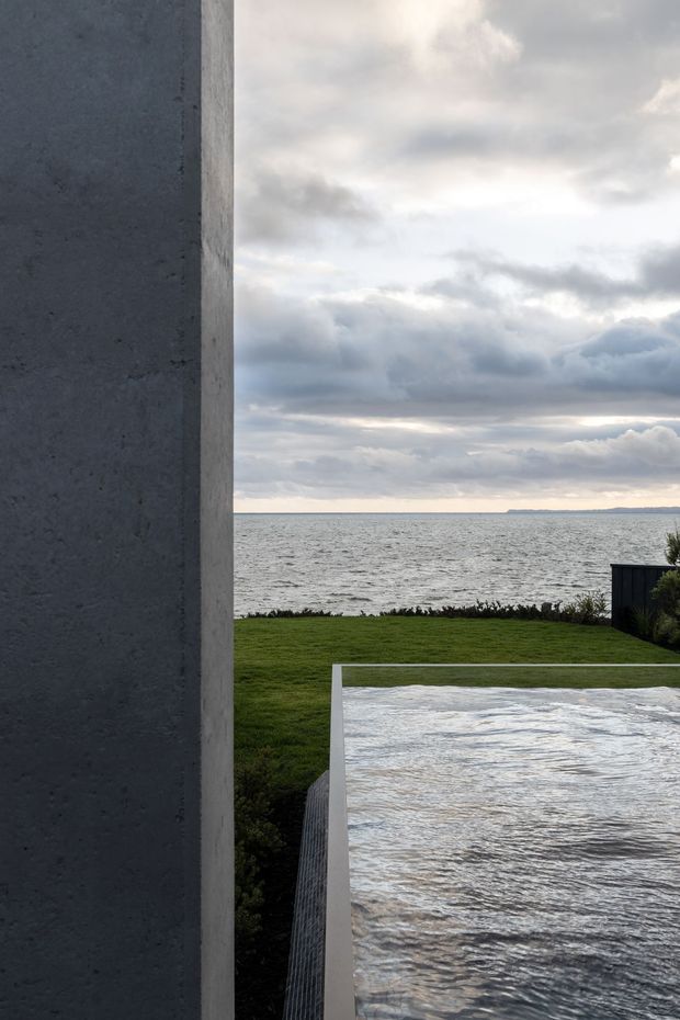 A minimalist coastal exterior view framed by a tall dark grey concrete wall on the left. A reflective rectangular pool or water feature occupies the foreground, with a neatly maintained lawn and low planting leading to a broad ocean horizon beneath an overcast sky.