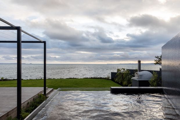 A modern coastal outdoor entertaining area overlooking the ocean, featuring a shallow reflective pool or water feature in the foreground, a timber deck, dark metal pergola structure, manicured lawn, and a built-in outdoor fireplace or barbecue beside a textured exterior wall. The scene has an overcast, dramatic sky and a calm waterfront setting.