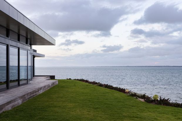 Modern waterfront house exterior with broad floor-to-ceiling glazing, projecting flat roof planes, and a timber-clad terrace overlooking a calm body of water. A manicured lawn and low coastal planting occupy the foreground beneath an overcast sky.