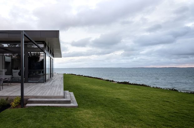 A modern glass-fronted coastal house with a flat roof and dark slender framing, opening onto a broad timber deck and steps. The building sits directly beside a grassy waterfront edge, with expansive views across the water under an overcast sky.