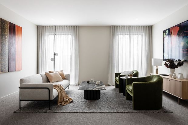 A refined contemporary living room with a neutral palette, soft grey carpet, a light upholstered sofa, sculptural black coffee table, and deep green lounge chairs. Floor-to-ceiling sheer curtains frame two windows, while abstract wall art and a timber-toned sideboard add colour and warmth. The room has a clean, balanced composition and generous natural light.